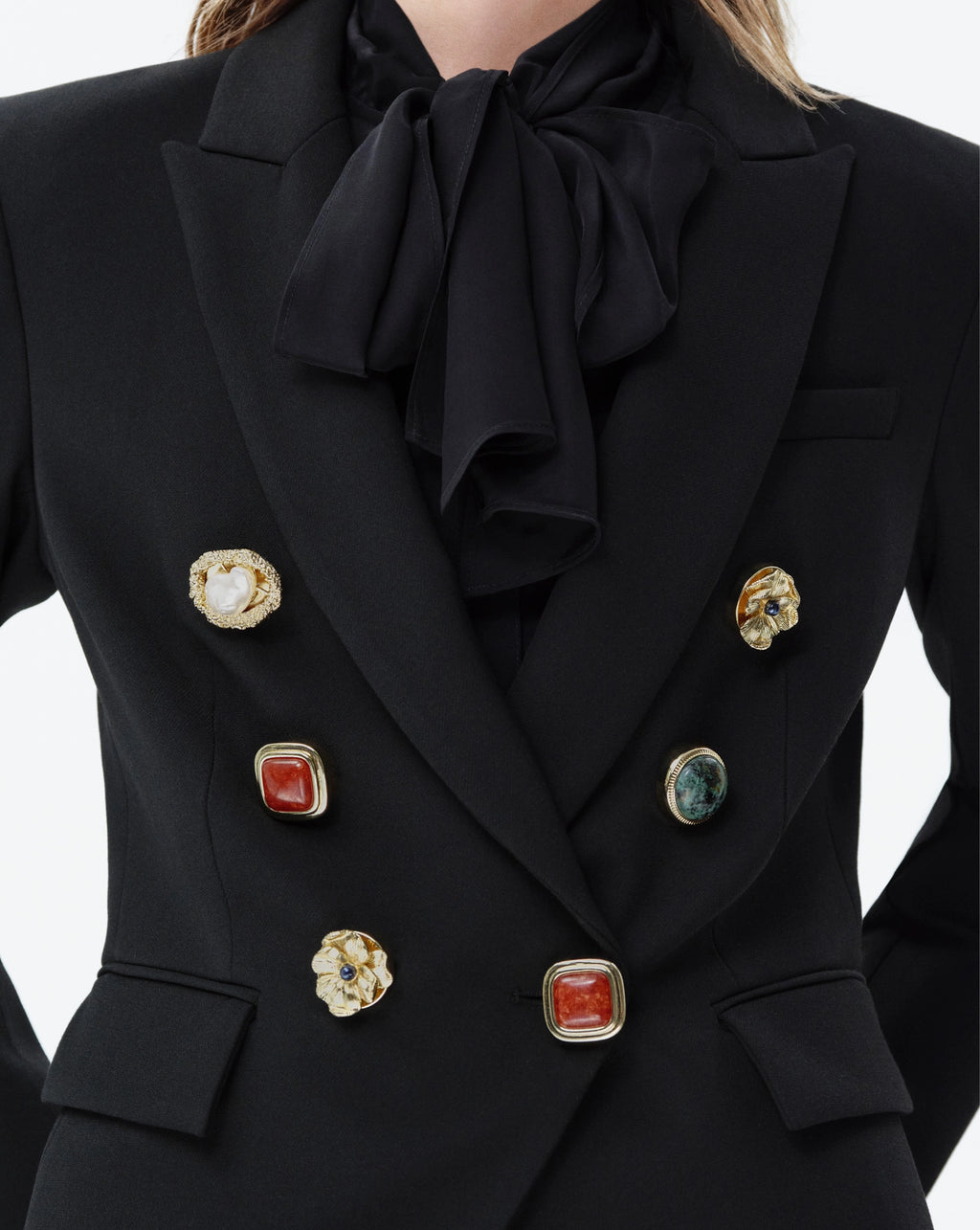 Close-up of a black double-breasted blazer with a large satin bow at the collar, featuring decorative gold-tone buttons with red, white, and green stone accents.