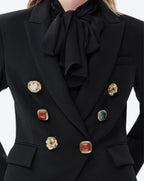 Close-up of a black double-breasted blazer with a large satin bow at the collar, featuring decorative gold-tone buttons with red, white, and green stone accents.