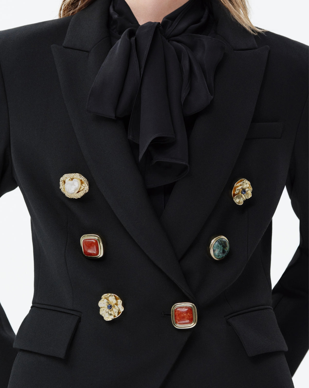 Close-up of a black double-breasted blazer with a large satin bow at the collar, featuring decorative gold-tone buttons with red, white, and green stone accents.