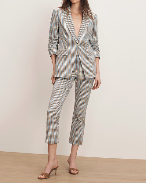 A woman wearing a checkered suit stands in a minimalist setting, her expression neutral as she poses for the camera.