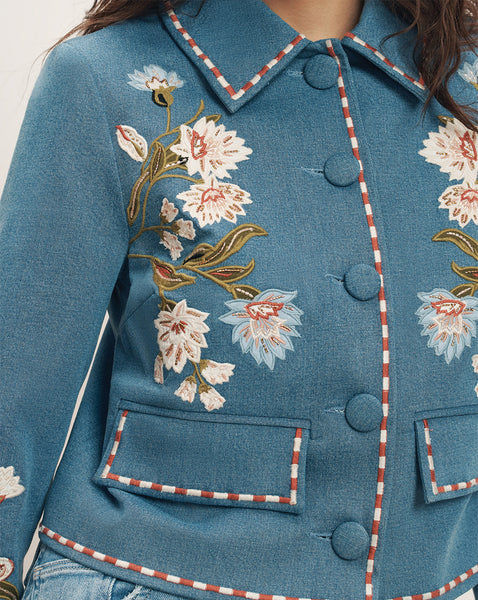 A close-up view of a woman's upper body, showcasing a beautifully embroidered floral pattern on a teal-colored jacket with decorative buttons and trim.