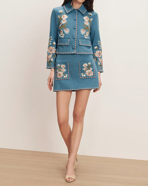 A woman wearing a teal-colored floral embroidered dress is standing on a wooden floor against a plain background.