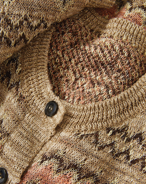 The image depicts a close-up view of a textured, knitted or crocheted fabric, with a button visible in the foreground. The background appears to be a natural, earthy-toned surface, possibly wood or a similar material.