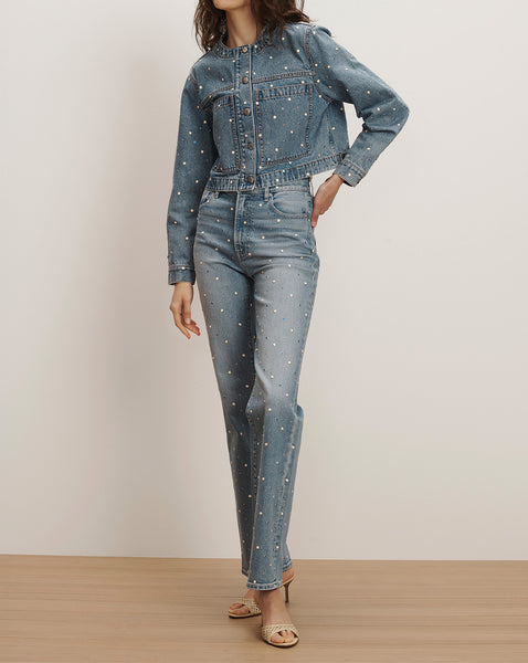 The image shows a woman wearing a denim jumpsuit with white polka dots, standing in a plain room with a wooden floor.