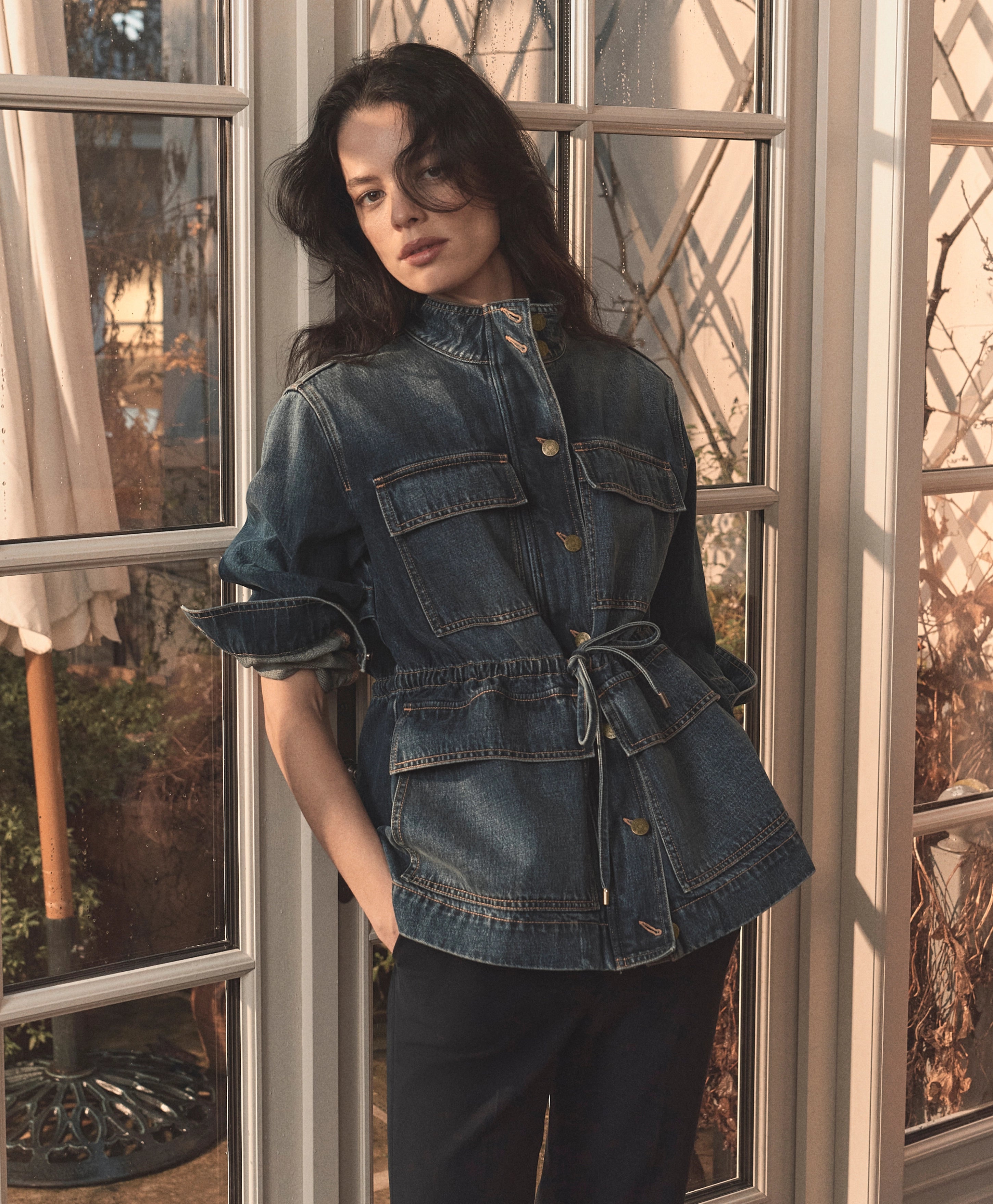 A young woman with dark curly hair stands in front of a window, wearing a denim jacket and looking directly at the camera.