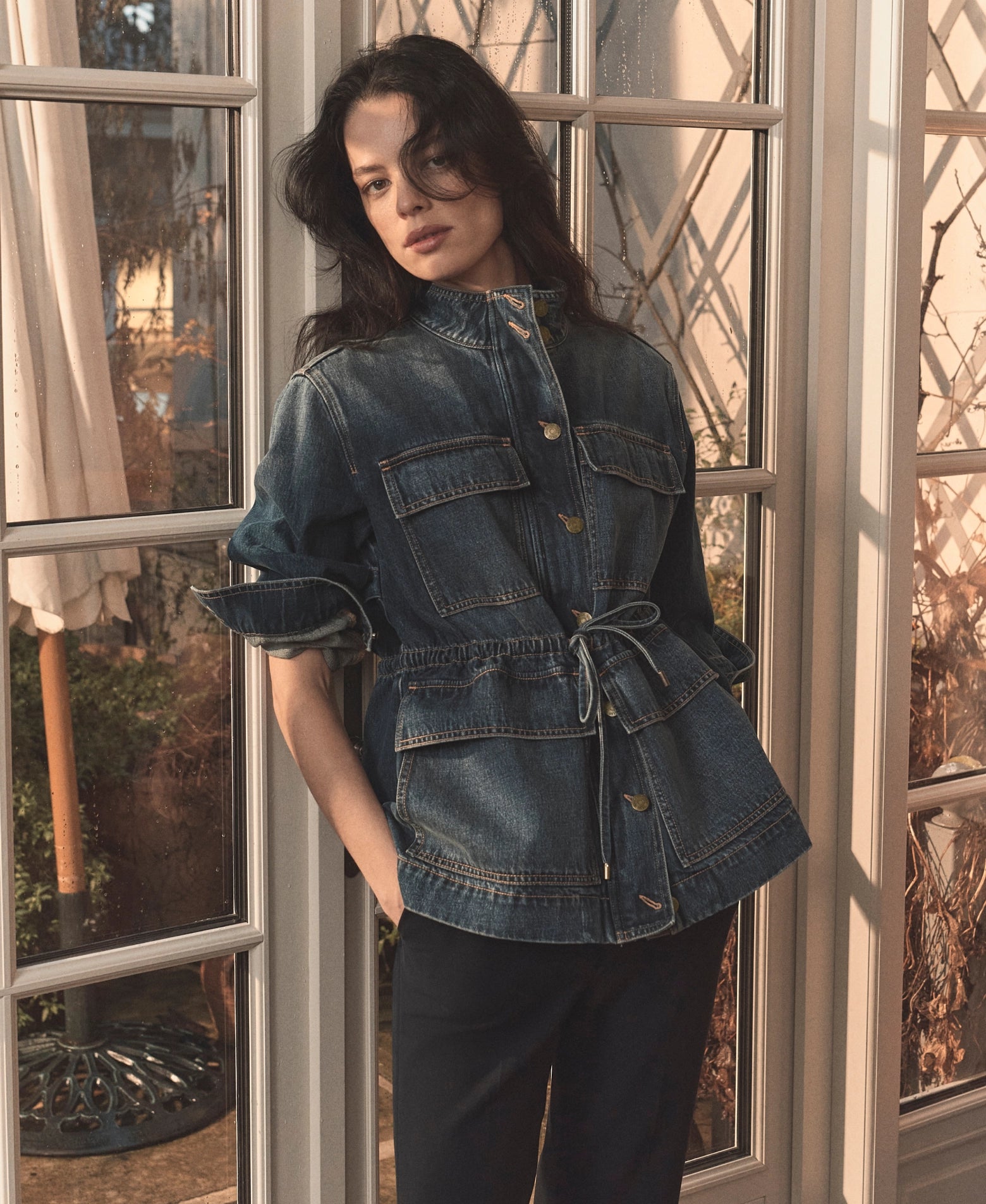 A woman with dark hair stands in front of a window, wearing a denim jacket and looking thoughtfully out the window.