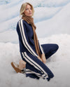 A woman with long blonde hair sits in the snow, wearing a navy blue and white striped sweater and scarf, with a serene expression on her face.