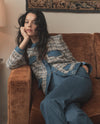 A young woman with long dark hair is sitting on a couch, wearing a patterned sweater and blue pants, and appears to be resting her head on her hand in a thoughtful pose. The background includes a decorative wall hanging and a wooden chair.
