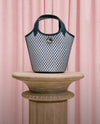 A black and white patterned handbag sits on a wooden pedestal against a pink curtain backdrop.
