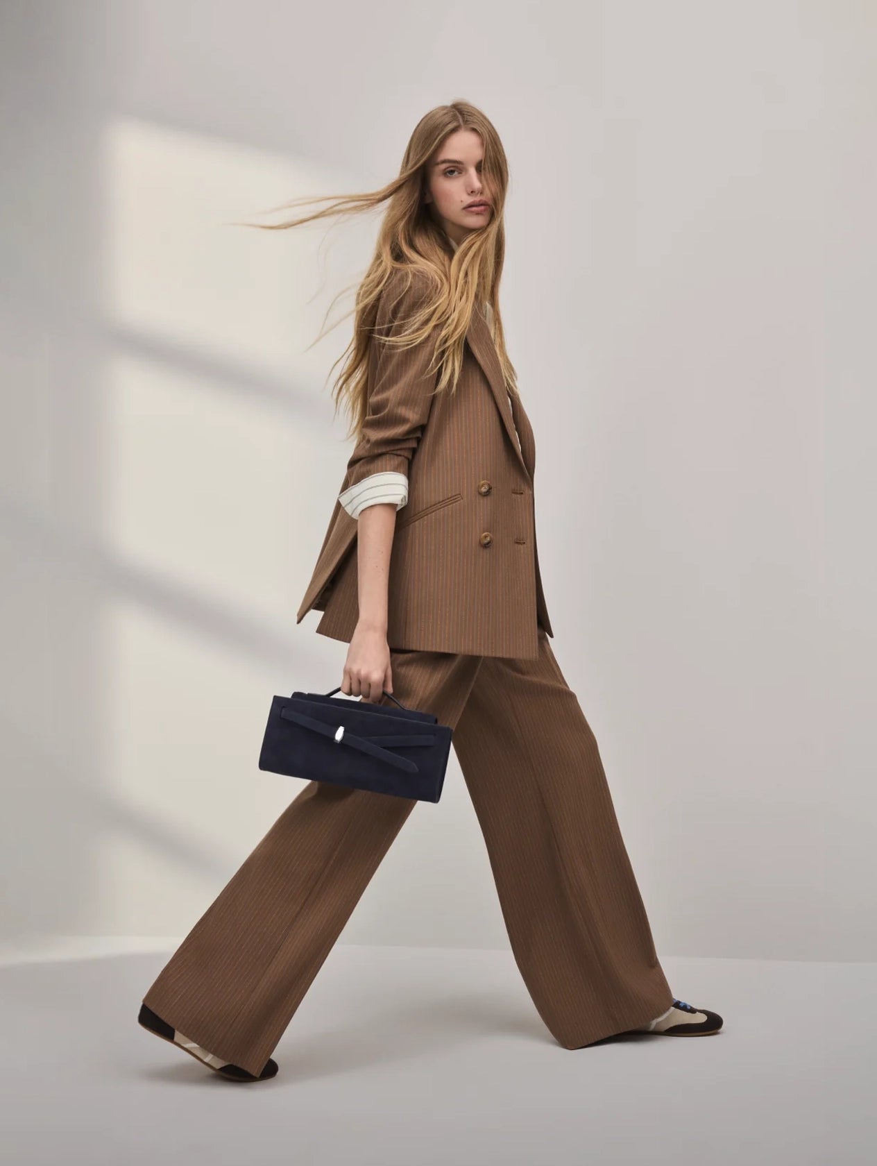 A young woman with long, windblown blonde hair is wearing a tan suit and carrying a navy blue briefcase as she walks across a plain white background.